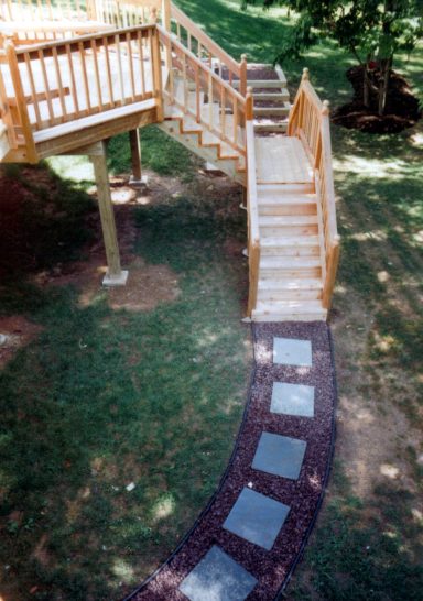 Wooden stairs leading up to a deck, with a stone path winding through green grass.