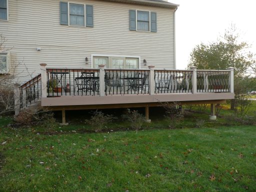 Wooden deck with tables and chairs, attached to a house, surrounded by grass and shrubs.