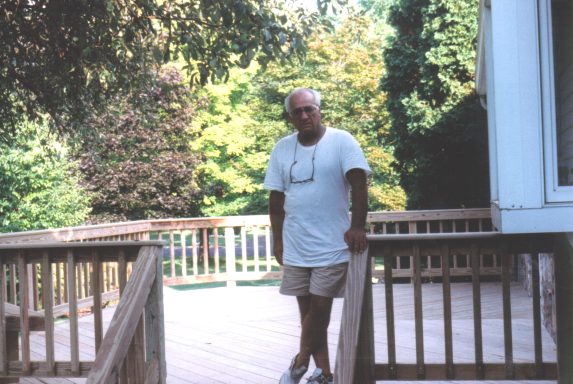 A man stands on a wooden deck surrounded by greenery.
